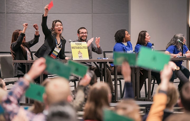 Individuals are seated on a raised platform behind long tables. One of the individuals is standing partway out of their chair and holding a red card above their head. In the foreground, you can see other colored index cards being held by audience members above their heads.