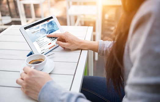 On a small rectangular table, a white coffee cup on a saucer and a tablet propped up with a stand are sitting next to each other. A woman's hand is holding the handle of the coffee cup while her other hand is touching the tablet screen. The tablet screen depicts the SOT Job Bank website. The woman's face is not visible in the photo.