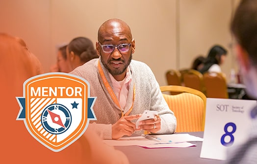 A black man sits at a table. He appears to be in conversation with unseen individuals at his table. Overlaid on the photo is a graphic of a orange and blue shield/badge that reads MENTOR.